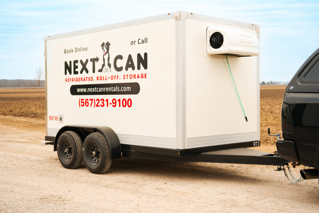 Side view of a white Next Can refrigerated trailer connected to a black pickup truck, parked in a vast open field.