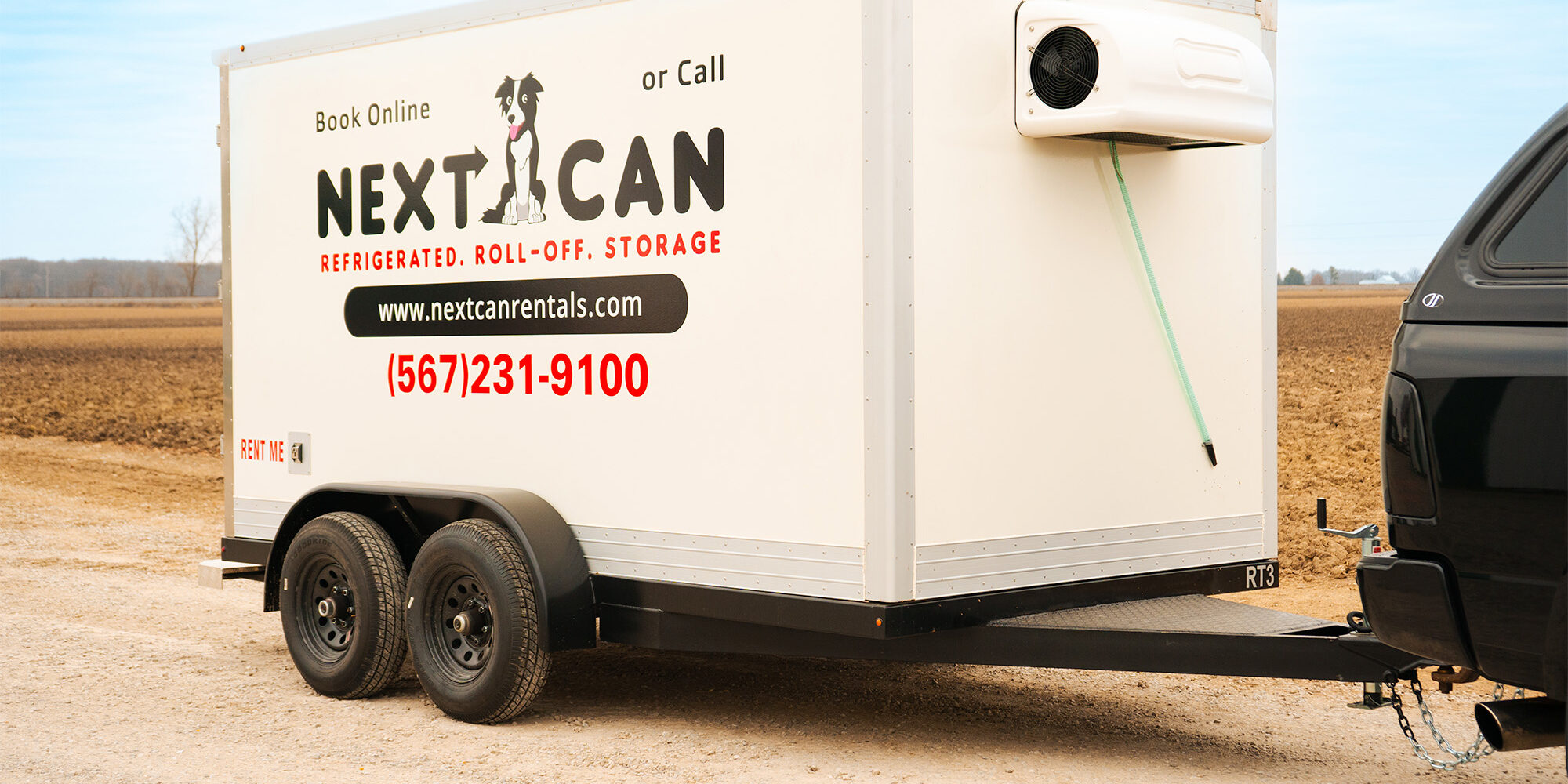 Side view of a white Next Can refrigerated trailer connected to a black pickup truck, parked in a vast open field.