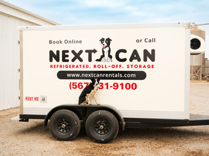 Side view of a white Next Can refrigerated trailer featuring the company logo and a dog mascot, parked in a rural setting.