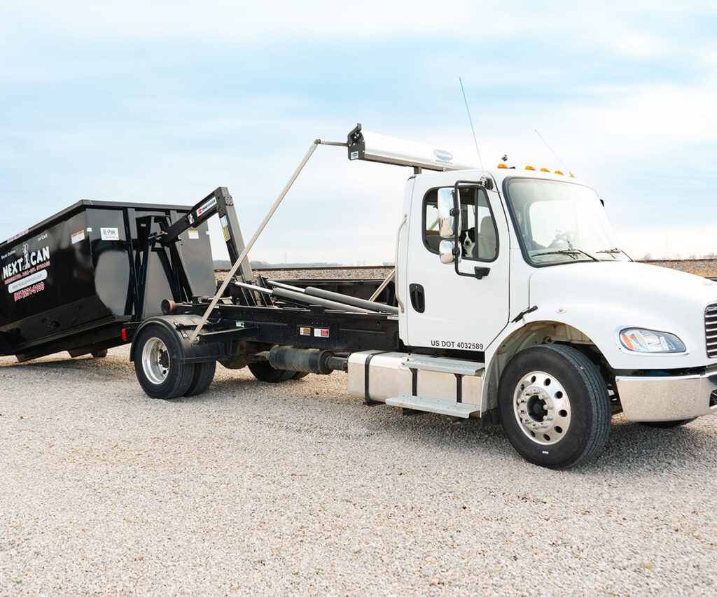 A white Next Can roll-off dumpster truck loading a black dumpster onto its bed in a gravel parking lot.