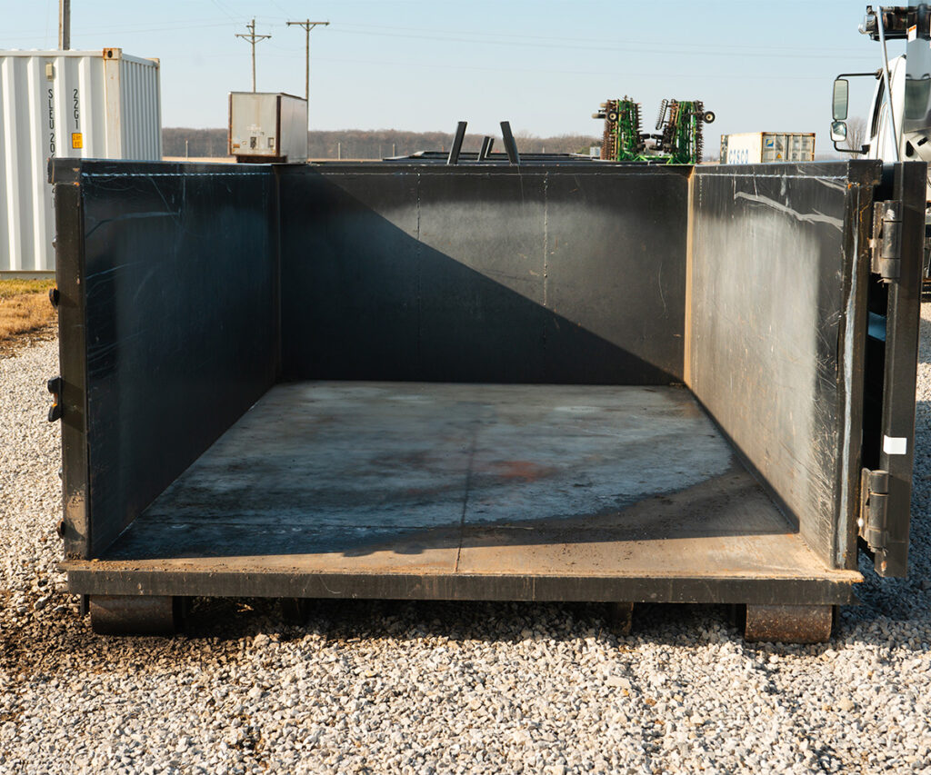 Broad side perspective of an empty, large black roll-off dumpster on a gravel ground, with industrial surroundings.